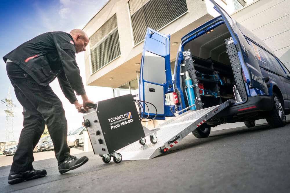 Ein Handwerker in Arbeitskleidung schiebt ein mobiles Schweißgerät über eine Rampe aus einem blauen Lieferwagen.