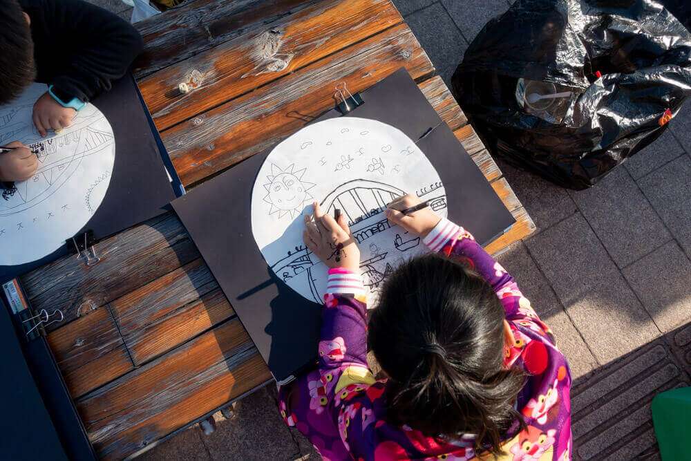 Zwei Kinder sitzen an einem Holztisch und zeichnen mit Stiften auf runden Papierbögen, die auf schwarzen Unterlagen befestigt sind.