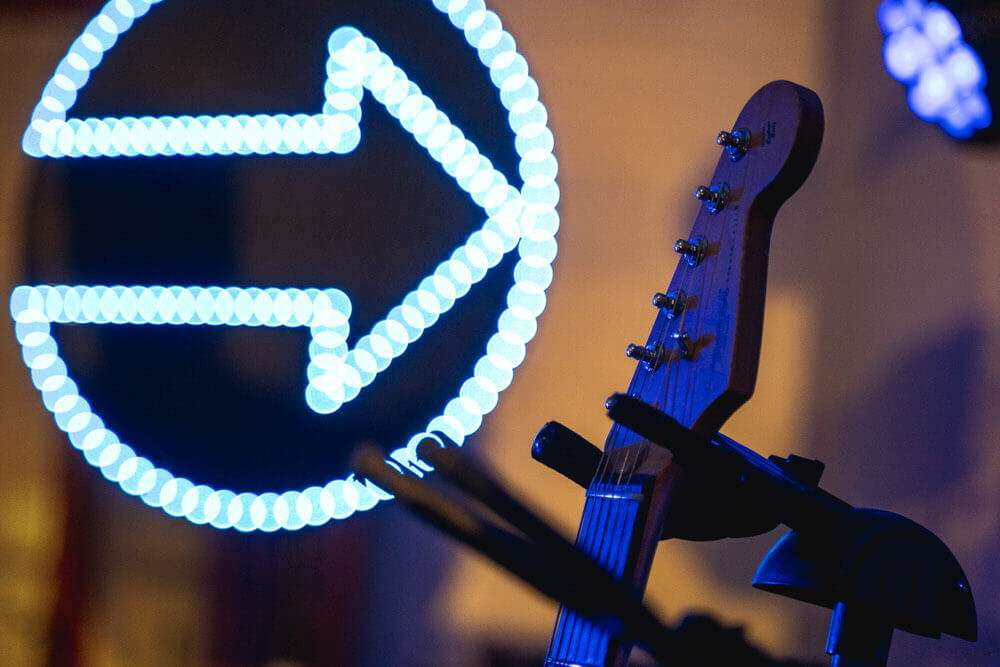 Der Hals einer E-Gitarre steht im Vordergrund, während im Hintergrund ein leuchtendes, blaues Pfeilsymbol in einem Kreis unscharf zu sehen ist.