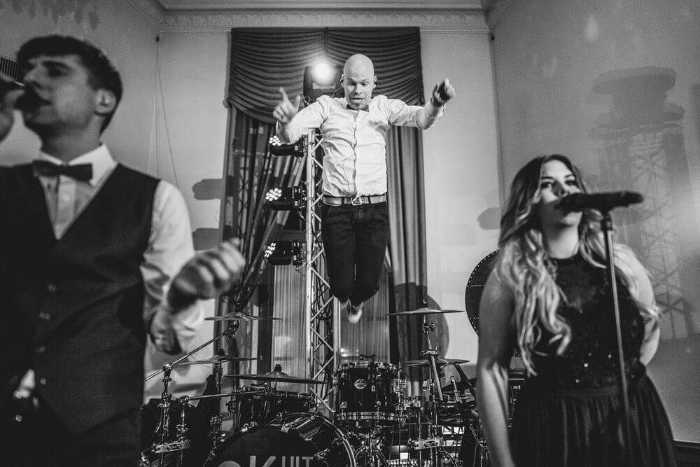 Eine Musikband tritt auf einer Bühne auf, wobei der Schlagzeuger in der Luft springt, während eine Sängerin und ein weiterer Musiker im Vordergrund zu sehen sind.