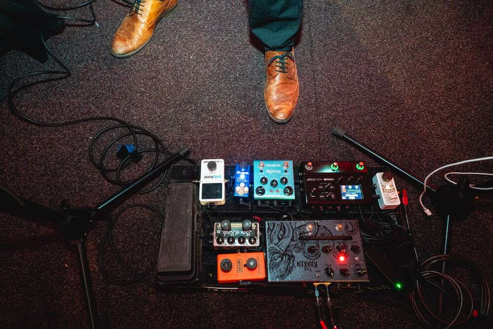 Ein Paar braune Lederschuhe steht vor einem Effektpedalboard für Gitarren auf einem dunklen Bühnenboden.