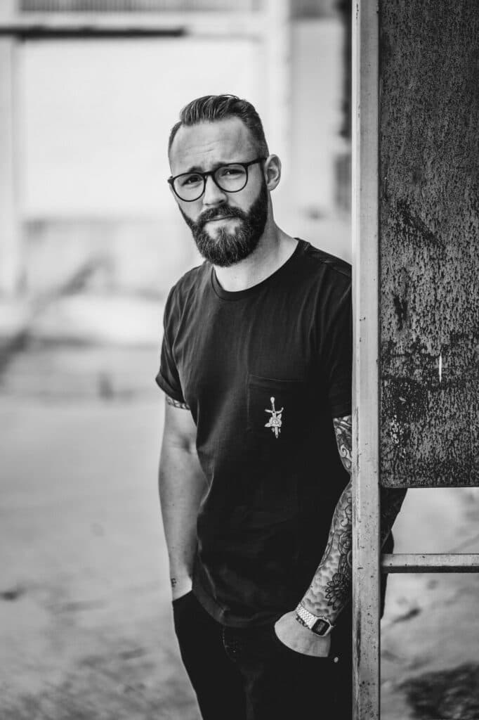 Ein bärtiger Mann mit Brille und tätowierten Armen steht in einem T-Shirt an einem metallenen Pfosten und blickt direkt in die Kamera.