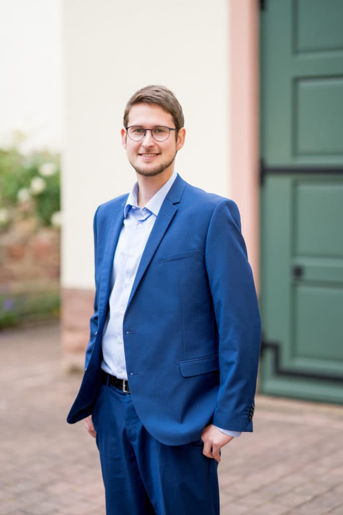 Ein junger Mann mit Brille in einem blauen Anzug und hellem Hemd steht lächelnd vor einer grünen Tür und einer hellen Fassade.