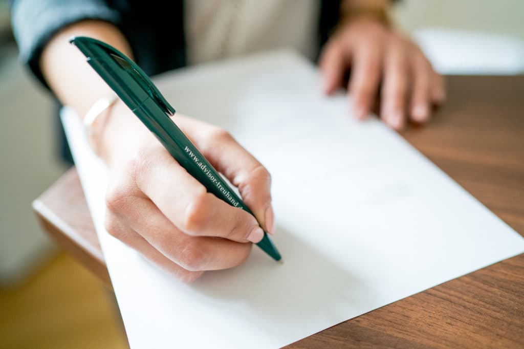 Nahaufnahme einer Hand, die mit einem grünen Stift auf einem weißen Blatt Papier schreibt, das auf einem Holztisch liegt.