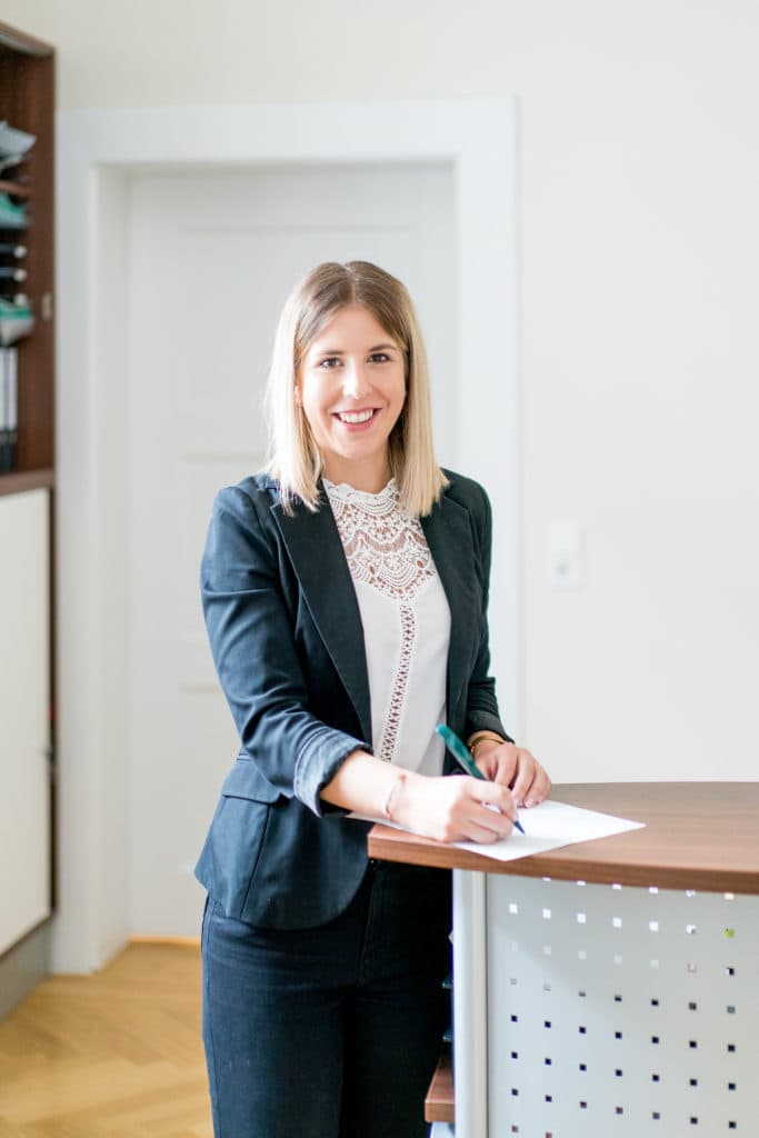 Eine blonde Frau in einem dunklen Blazer und einer weißen Spitzenbluse steht an einem Stehpult und schreibt auf einem Blatt Papier, während sie in die Kamera lächelt.