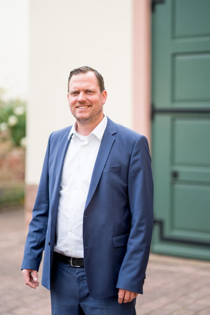 Ein lächelnder Mann in einem blauen Anzug und weißem Hemd steht vor einem grünen Tor.