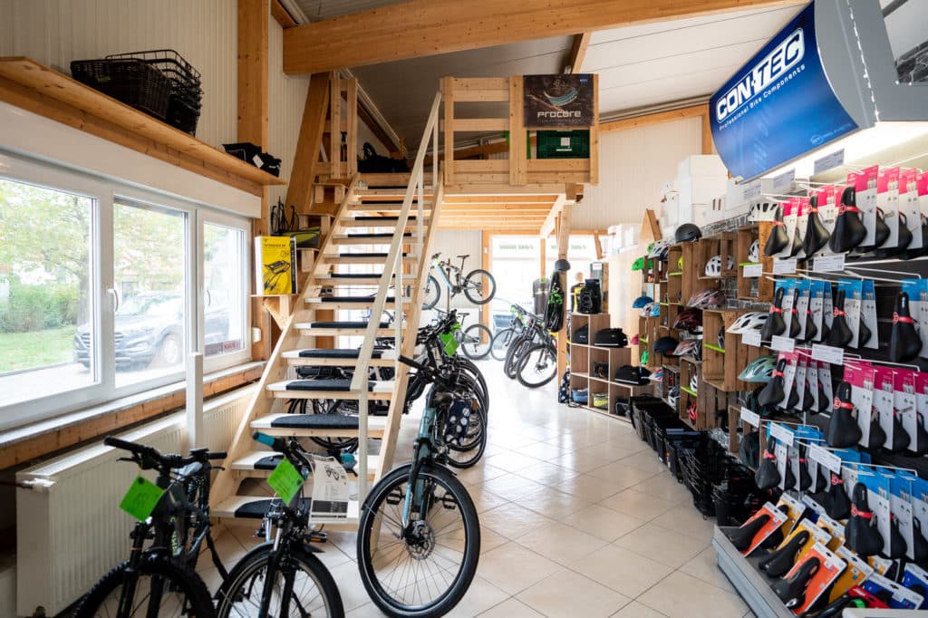 Blick in ein helles Fahrradgeschäft mit einer zentralen Holztreppe, Fahrrädern im Vordergrund und Regalen voller Zubehör an der rechten Wand.