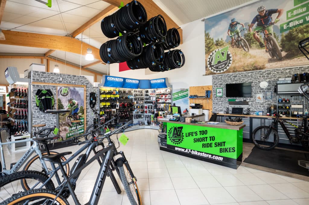 Blick in ein helles Fahrradgeschäft mit E-Bikes im Vordergrund, Reifen an der Decke und einer grünen Verkaufstheke.