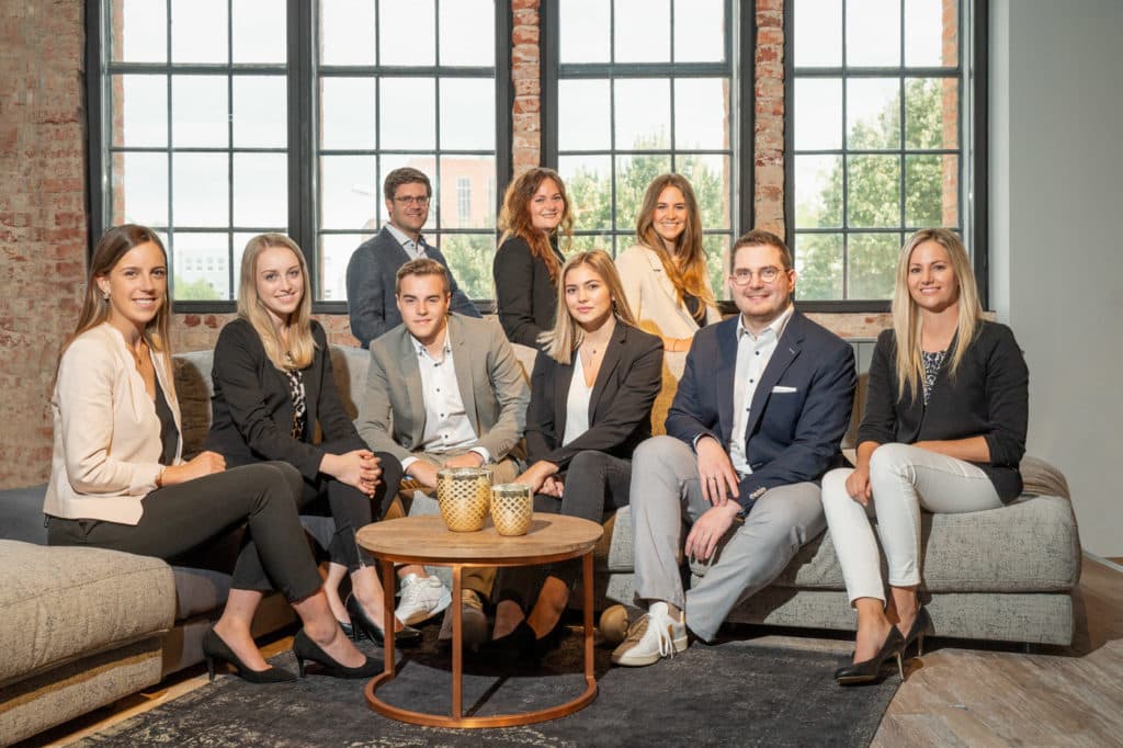 Eine Gruppe von acht Personen sitzt und steht in einem hellen Büro vor großen Fenstern auf einem Sofa und posiert für ein Foto.
