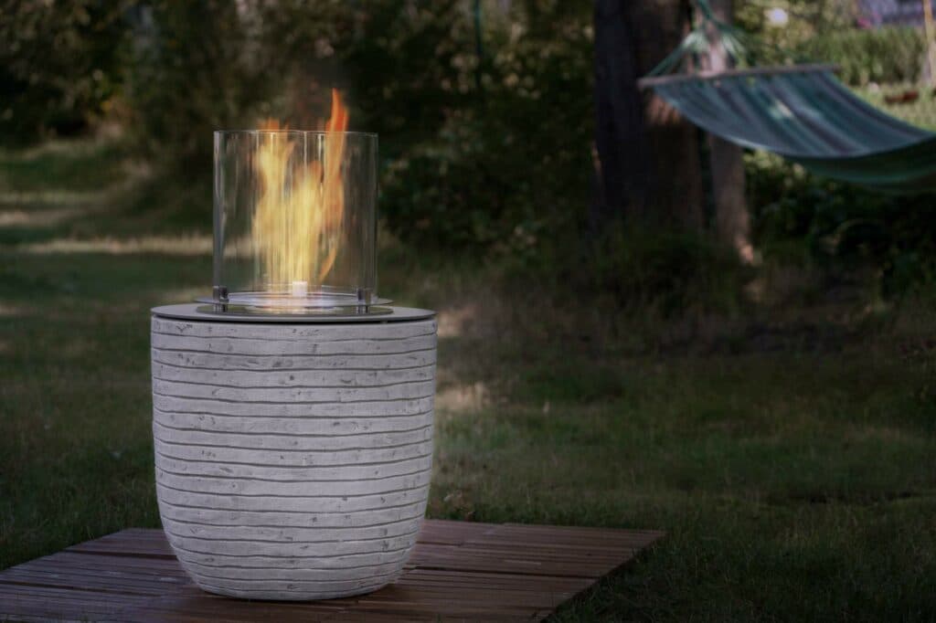 Ein runder, weißer Tischkamin mit brennender Flamme steht auf einer Holzplattform im Garten, im Hintergrund ist eine Hängematte zu sehen.