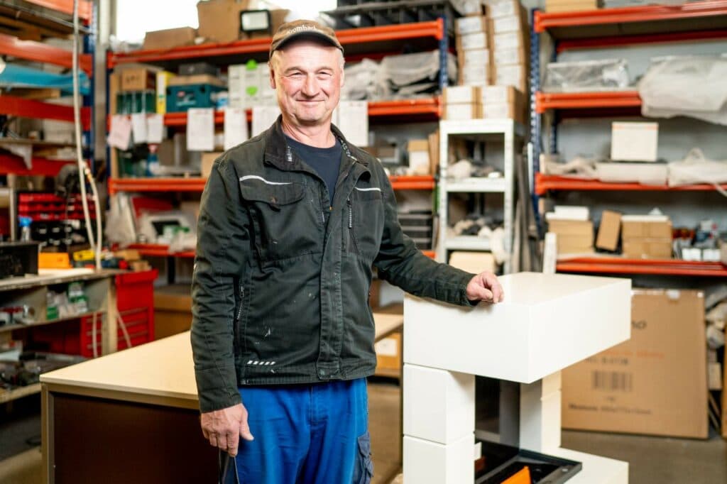 Ein Mann mit Kappe und dunkler Arbeitsjacke steht in einem Lagerraum vor einem weißen, modularen Möbelstück und lächelt in die Kamera.