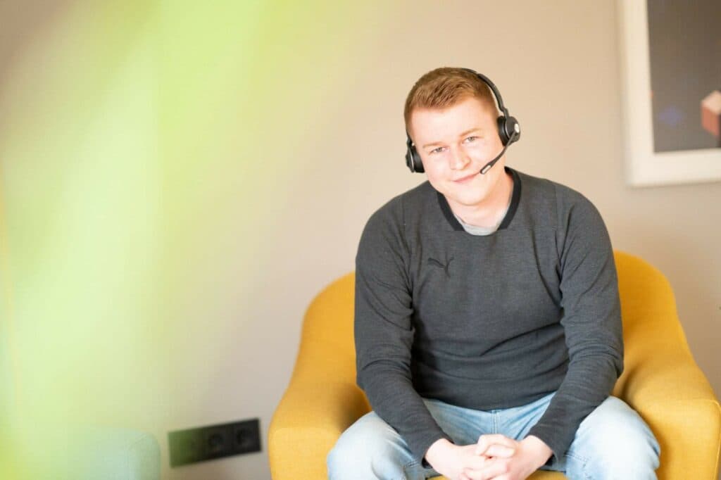 Ein junger Mann mit Headset sitzt in einem gelben Sessel vor einer hellen Wand.