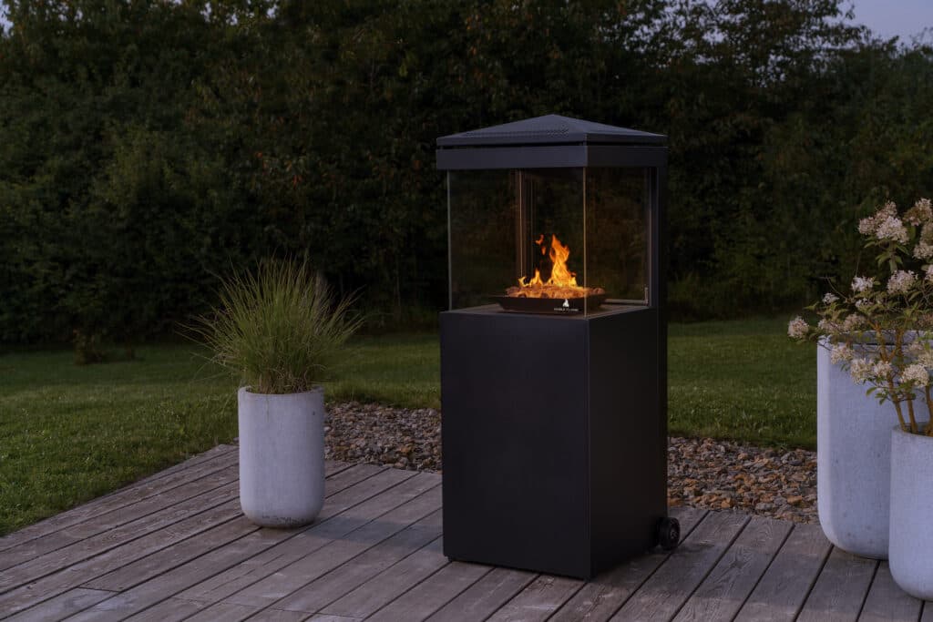 Ein schwarzer, moderner Terrassenkamin mit einer brennenden Flamme steht auf einer Holzterrasse vor einer grünen Hecke.