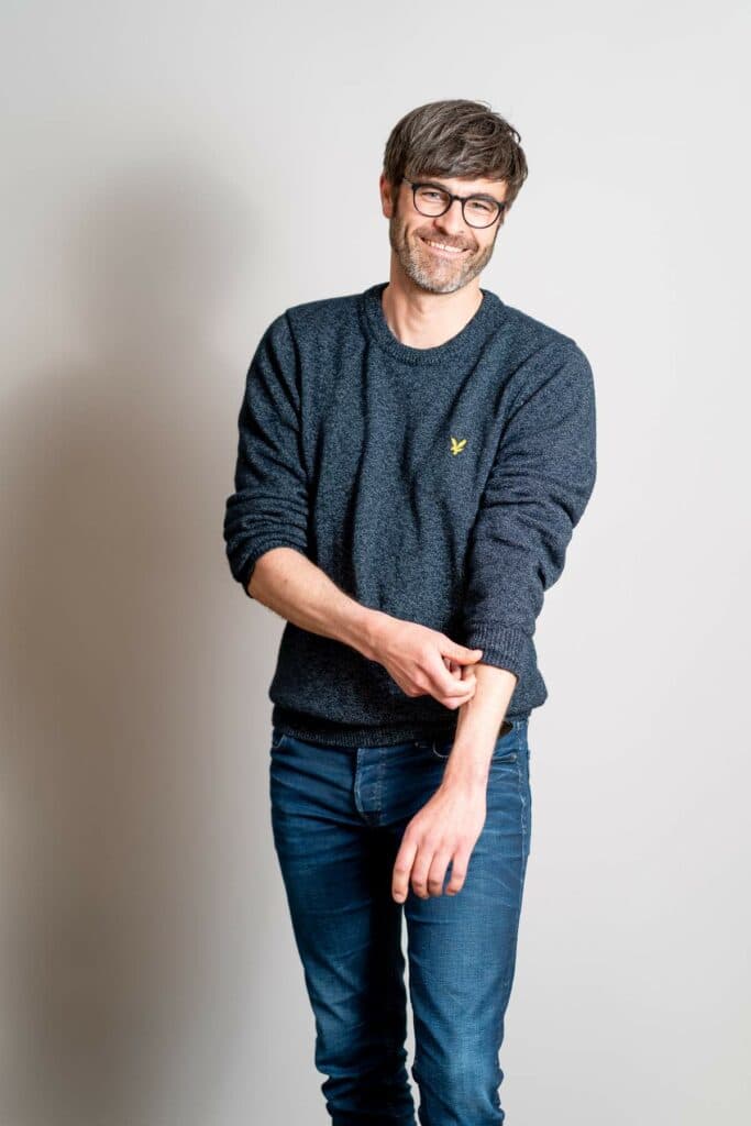 Ein lächelnder Mann mit Brille und Bart trägt einen dunkelblauen Pullover und Jeans vor einem hellgrauen Hintergrund.