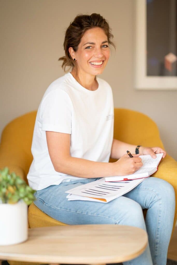 Eine lächelnde Frau in einem weißen T-Shirt und blauer Jeans sitzt in einem gelben Sessel und schreibt mit einem Stift in ein aufgeschlagenes Notizbuch.