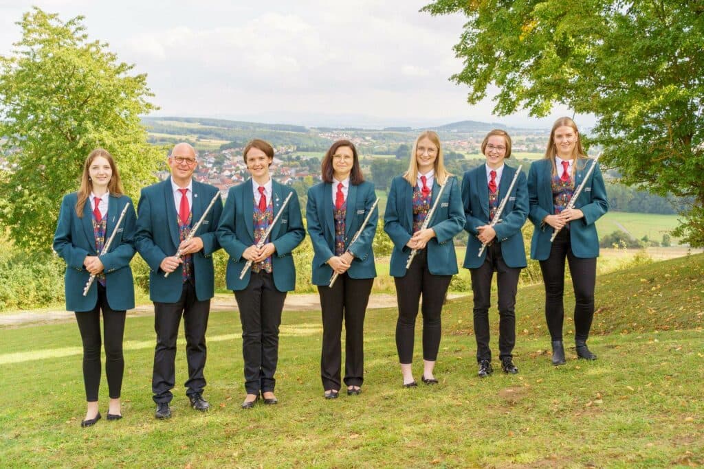 Eine Gruppe von sieben Musikern in einheitlicher Kleidung steht auf einer Wiese und hält Querflöten in den Händen.