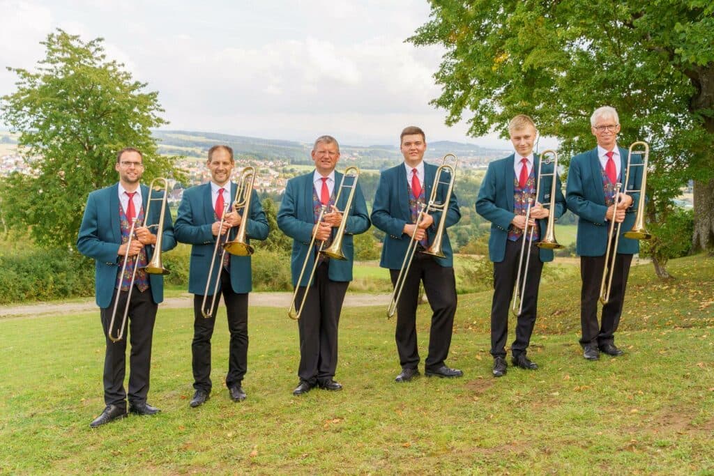Sechs Männer in blauen Blazern und roten Krawatten stehen nebeneinander auf einer Wiese und halten jeweils eine Posaune in der Hand.