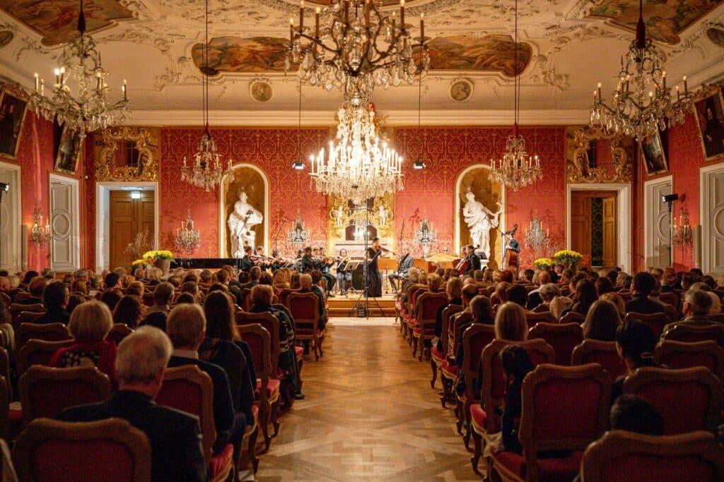 Blick in einen prunkvollen, rot ausgeschlagenen Festsaal mit Kronleuchtern, in dem ein Orchester vor einem Publikum spielt.