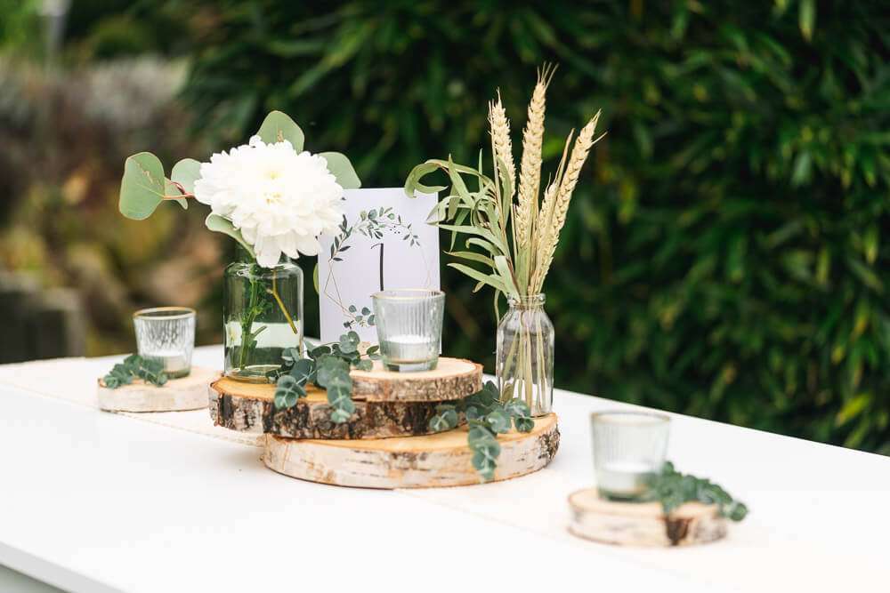 Eine festliche Tischdekoration mit weißen Blumen, Weizenähren, Eukalyptuszweigen und Kerzengläsern auf runden Holzscheiben vor einem unscharfen grünen Hintergrund.