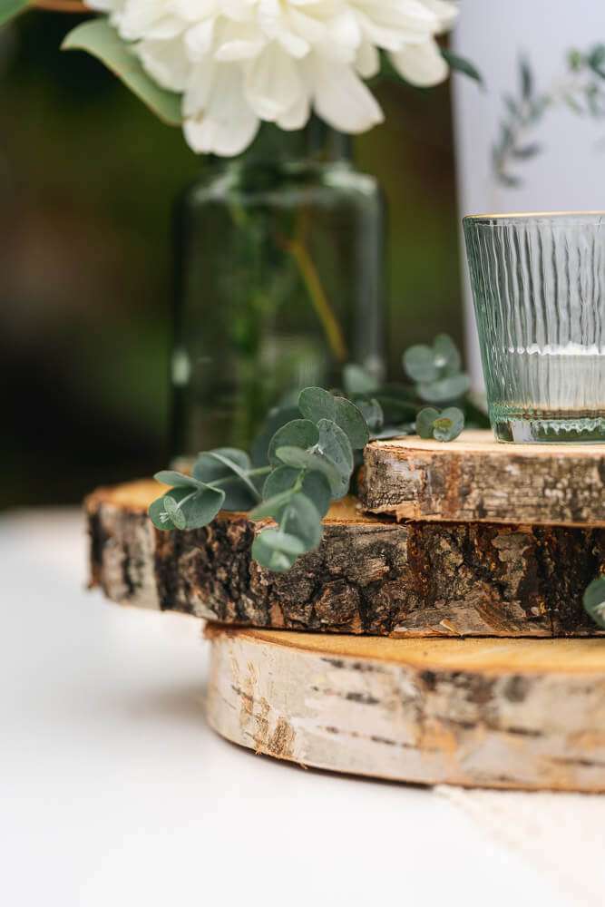 Nahaufnahme von drei übereinander gestapelten Holzscheiben auf einem Tisch, dekoriert mit einem Eukalyptuszweig, einer Glasvase mit weißen Blumen und einem gerippten Glas.