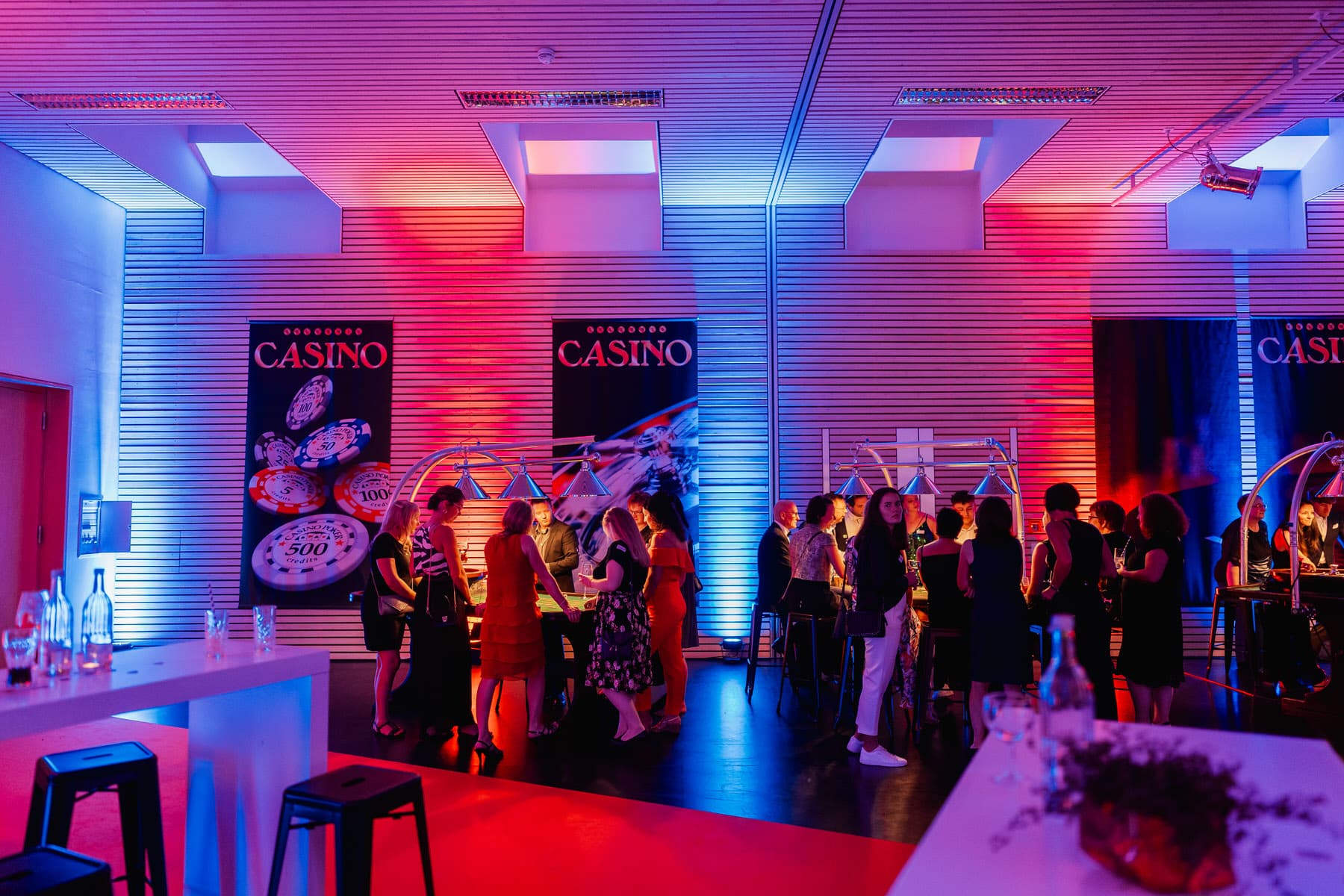 Ein Innenraum mit Casino-Thematik ist in blaues und rotes Licht getaucht, während sich eine Gruppe von Menschen an Spieltischen aufhält.
