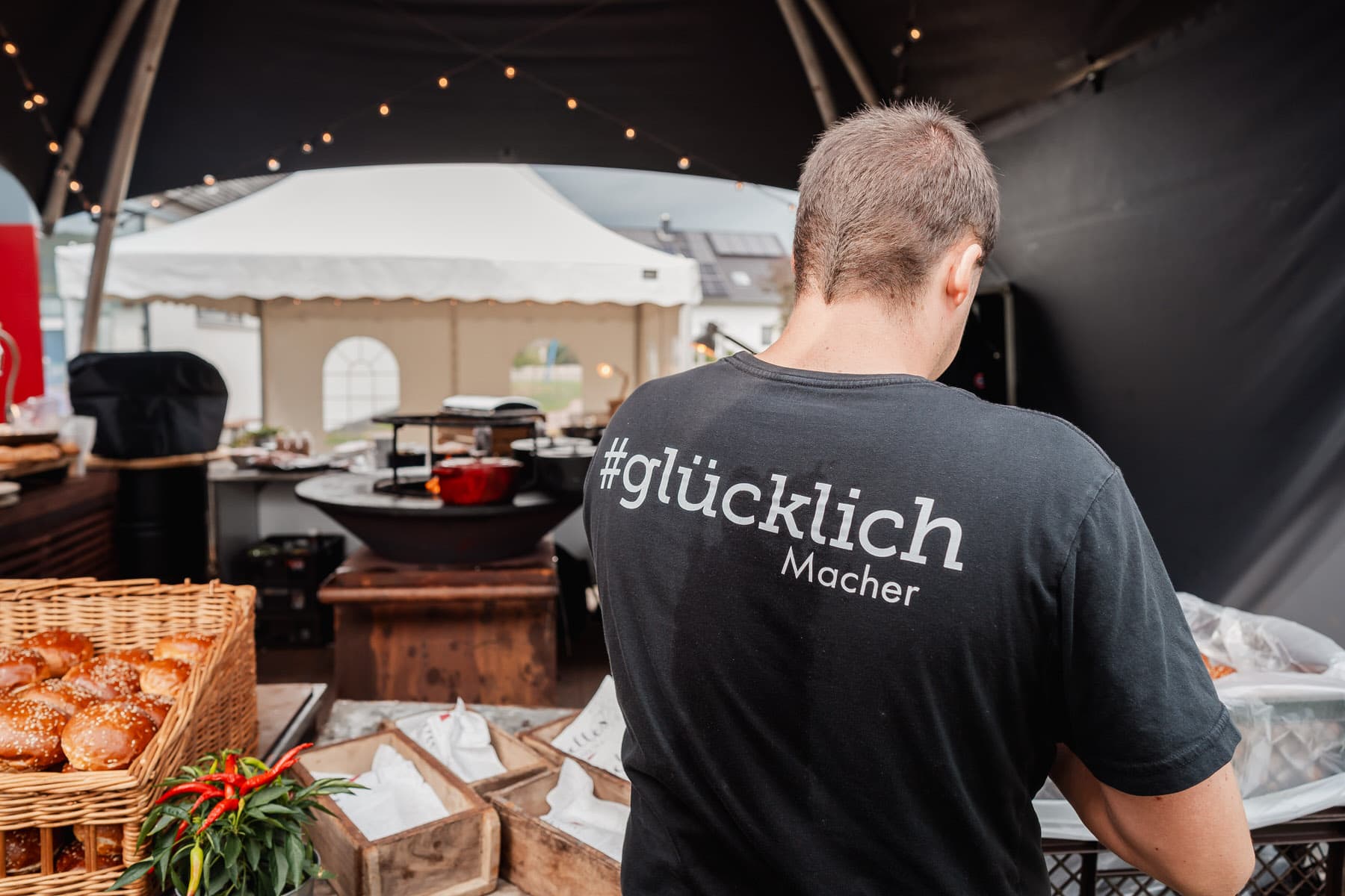Eine Person von hinten in einem schwarzen T-Shirt mit der Aufschrift glücklich Macher steht an einem Marktstand mit Backwaren.