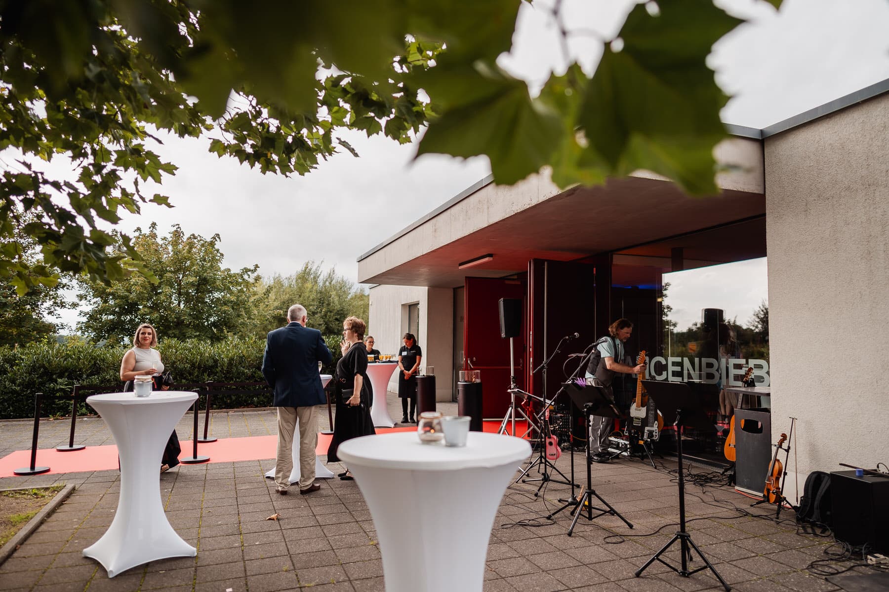 Menschen stehen an weißen Stehtischen vor einem modernen Gebäude, während im Hintergrund eine kleine Band spielt.