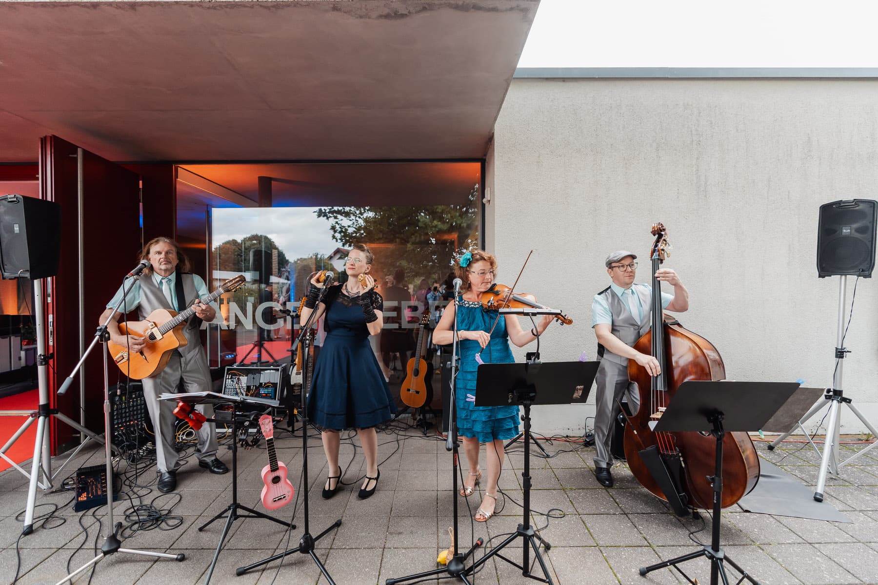 Eine vierköpfige Musikgruppe spielt auf einer Terrasse vor einem Gebäude Instrumente und singt in Mikrofone.