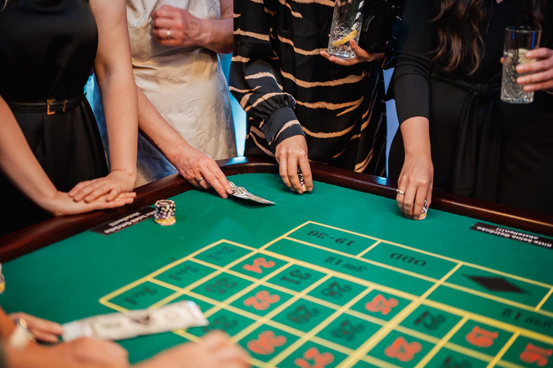 Mehrere Personen stehen um einen grünen Roulette-Tisch und legen ihre Hände auf das Spielfeld, während sie Gläser halten.