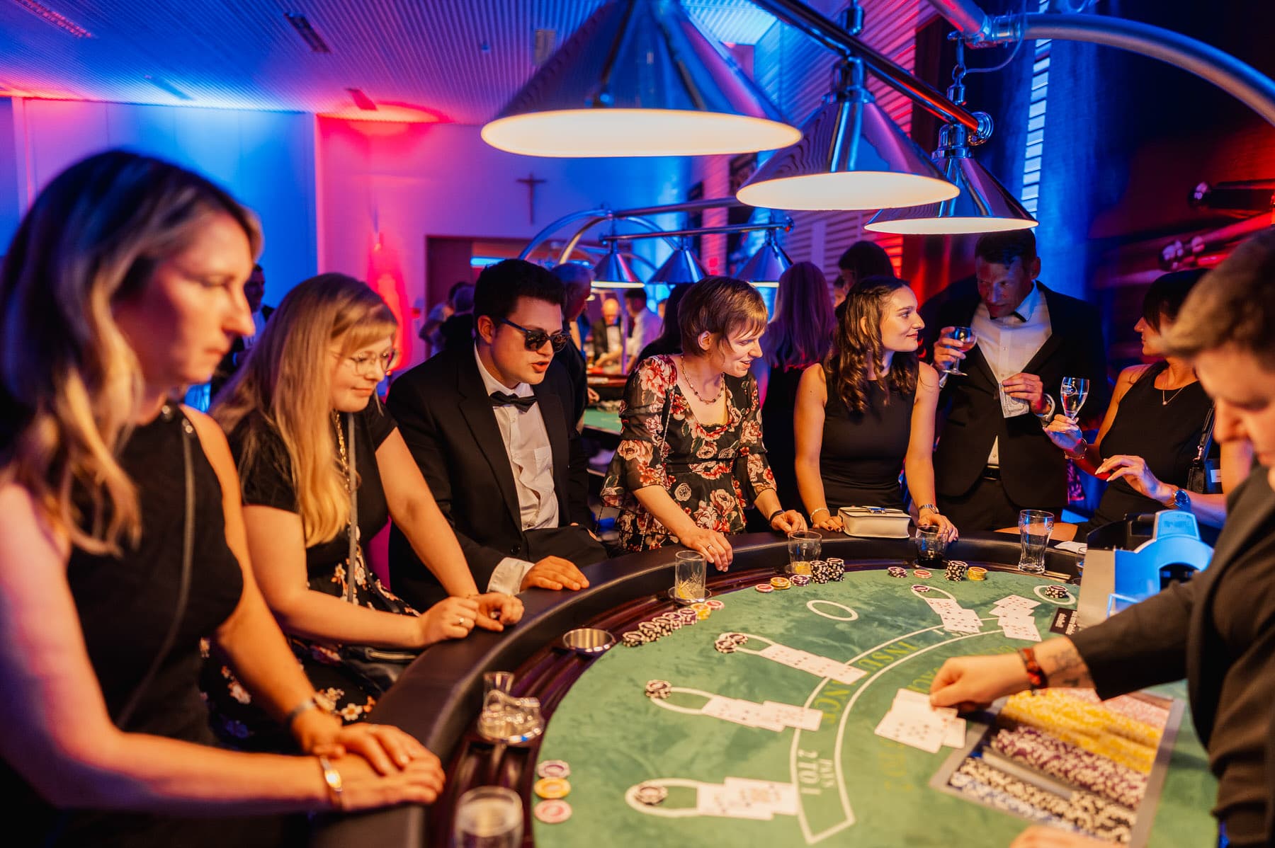 Eine Gruppe von Menschen sitzt an einem grünen Blackjack-Tisch in einem Casino mit blauer und roter Beleuchtung.