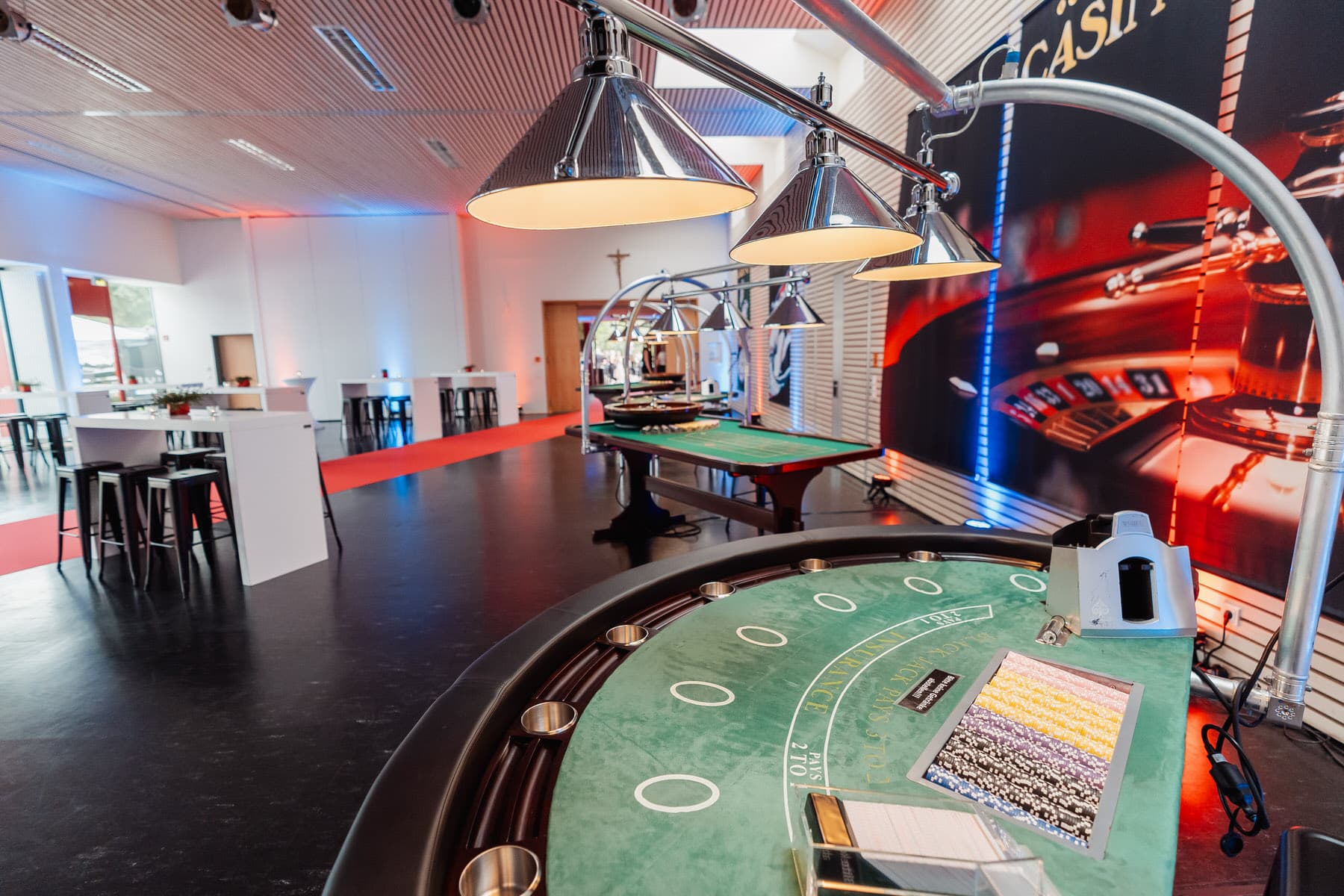 Ein Blick in einen modernen Casinoraum mit grünen Spieltischen, Barhockern und hellen Hängelampen.