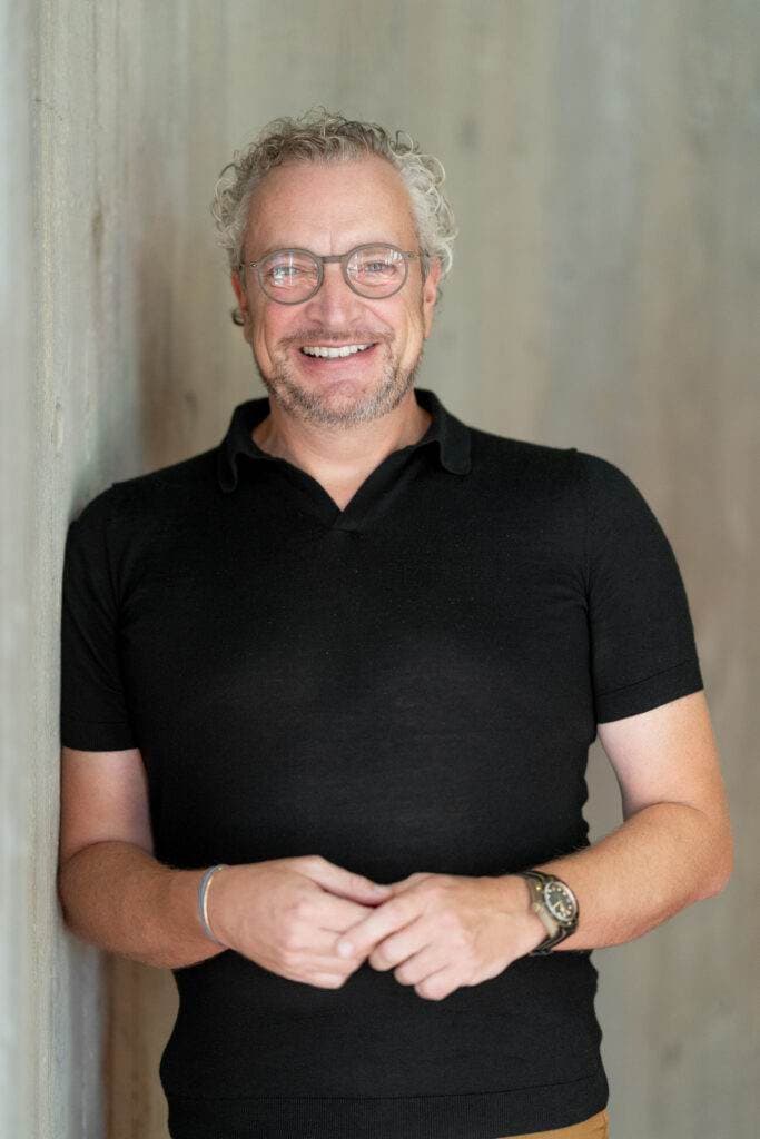 Ein lächelnder Mann mit Brille und schwarzem Polohemd steht vor einer grauen Betonwand.