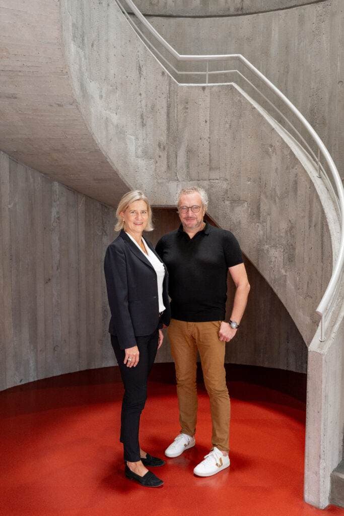 Ein Paar steht nebeneinander auf einem leuchtend roten Boden vor einer geschwungenen, grauen Betontreppe.