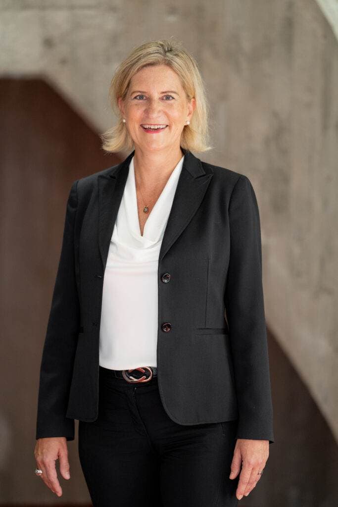 Eine blonde Frau mit einem weißen Oberteil und einem schwarzen Blazer lächelt in die Kamera, im Hintergrund ist eine graue Betonwand zu sehen.