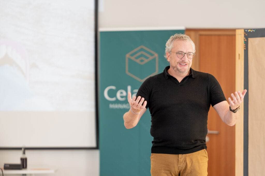 Ein Mann mit Brille und schwarzem Polohemd spricht vor einem grünen Firmenbanner und einer Projektionsfläche.