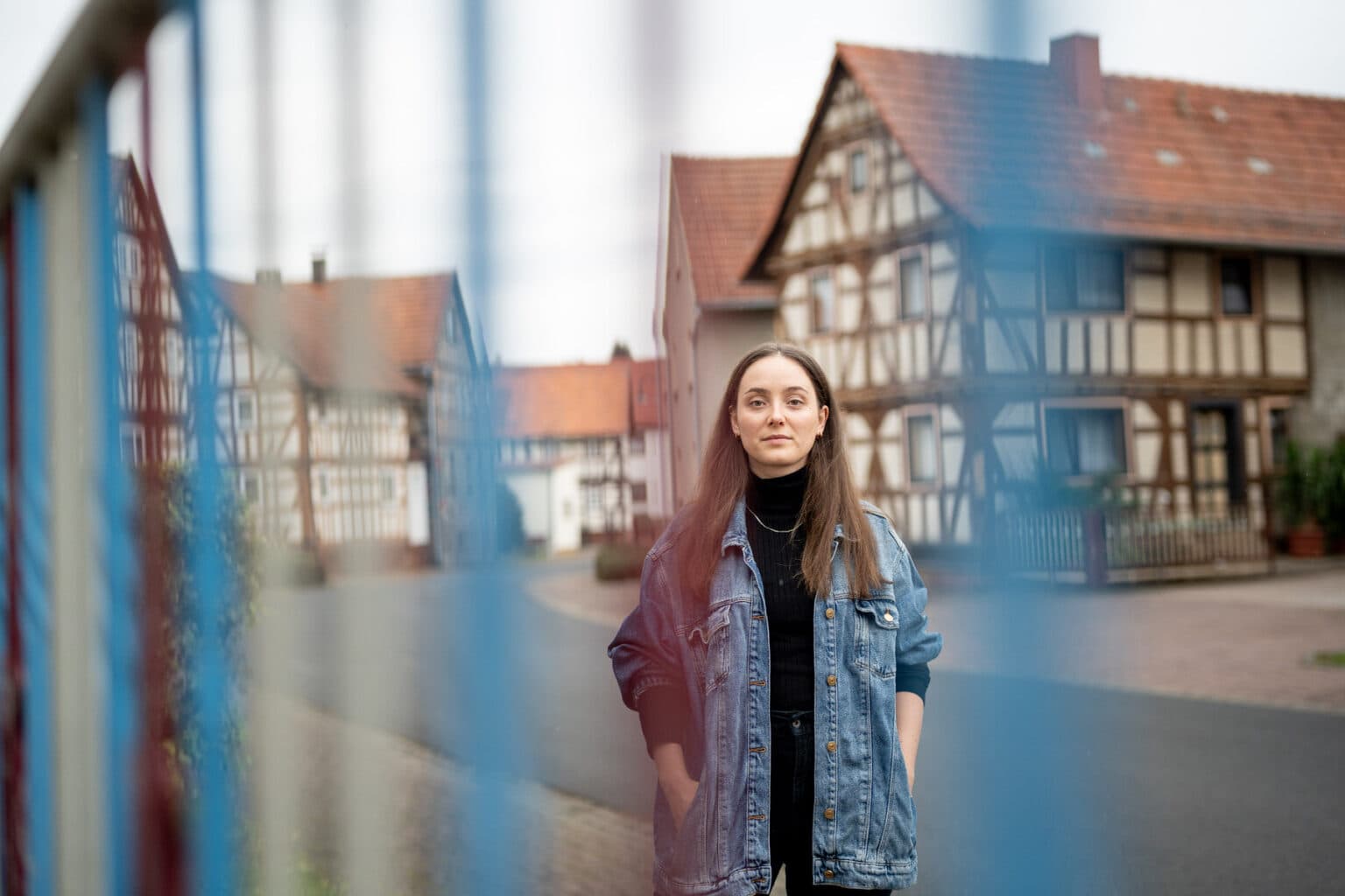 Eine junge Frau mit Jeansjacke steht auf einer Straße vor Fachwerkhäusern, wobei blaue Zaunstreben im unscharfen Vordergrund das Bild rahmen.