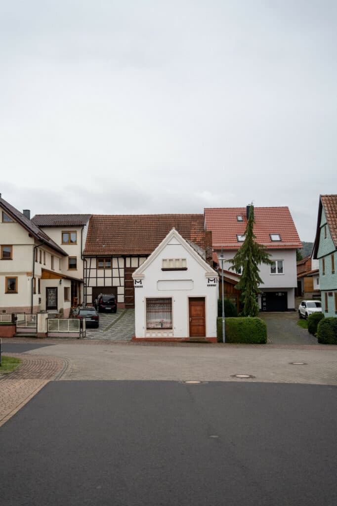 Blick auf eine Häuserzeile mit einem kleinen weißen Fachwerkhaus im Zentrum und einer asphaltierten Straße im Vordergrund unter bewölktem Himmel.