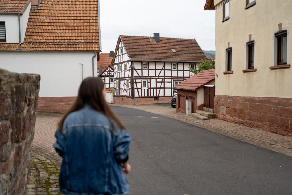 Eine Frau in einer Jeansjacke geht von hinten gesehen eine leere Straße in einem Dorf mit Fachwerkhäusern entlang.