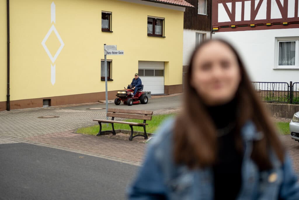Eine unscharfe Person im Vordergrund blickt in die Kamera, während im Hintergrund ein Mann auf einem kleinen Aufsitzmäher auf einer Straße vor einem gelben Haus fährt.