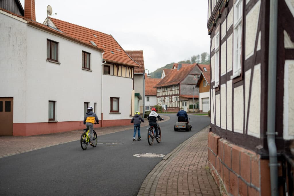 Eine Gruppe von Kindern und ein Erwachsener fahren auf einer asphaltierten Straße durch eine Ortschaft mit historischen Fachwerkhäusern.