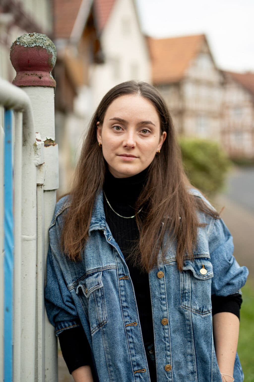 Eine junge Frau mit braunen Haaren trägt eine Jeansjacke über einem schwarzen Rollkragenpullover und steht vor einem unscharfen Fachwerkhaus.