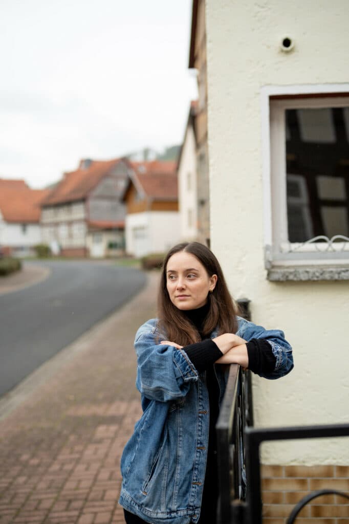 Eine junge Frau mit langen braunen Haaren trägt eine Jeansjacke und einen schwarzen Rollkragenpullover, während sie an einem schwarzen Metallgeländer vor einem hellen Fachwerkhaus steht.