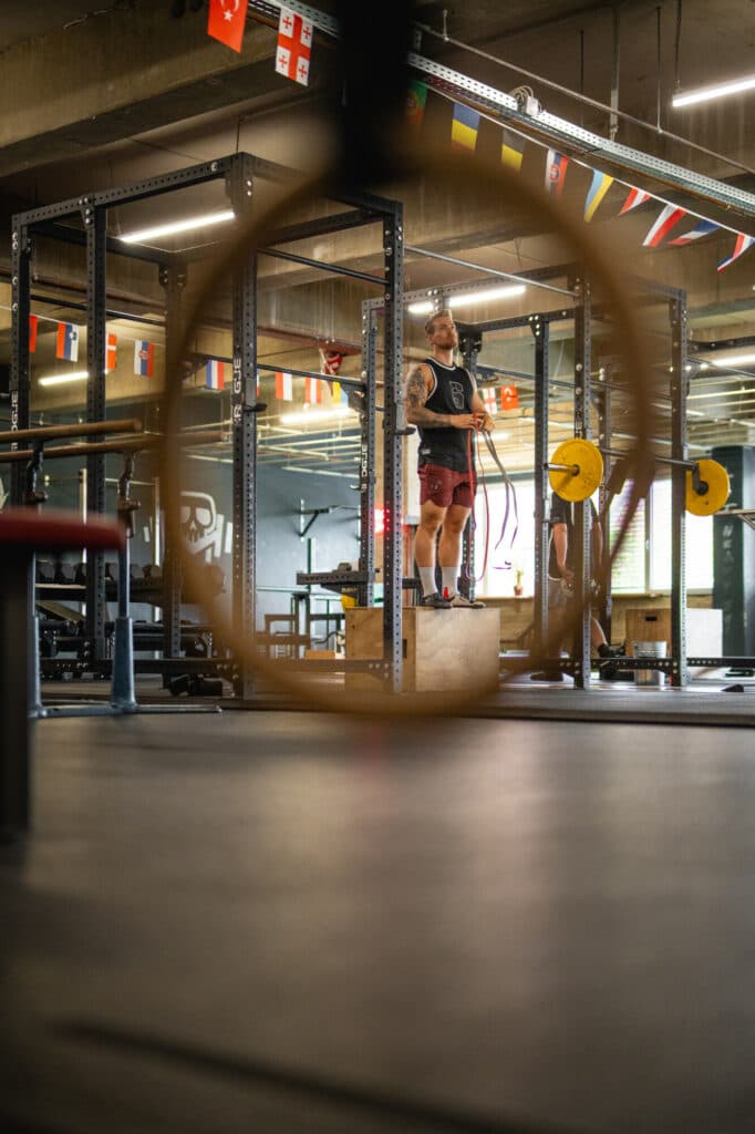 Ein Mann steht in einem Fitnessstudio auf einer Holzbox und hält Turnringe in den Händen, wobei der Fokus durch einen unscharfen Ring im Vordergrund auf ihn gelenkt wird.