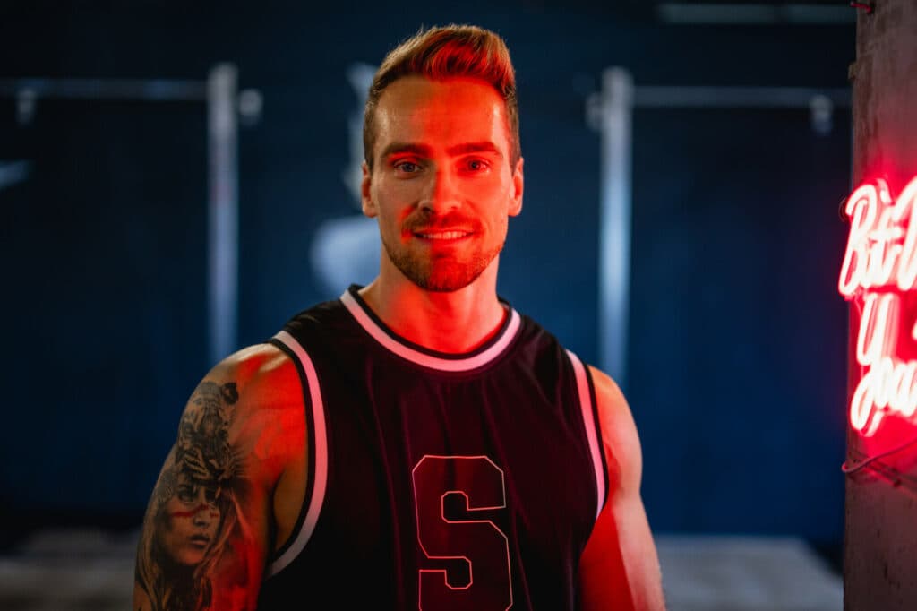 Ein junger Mann mit tätowiertem Oberarm trägt ein schwarzes Sporttrikot und lächelt in einem Fitnessstudio, das von rotem Neonlicht beleuchtet wird.