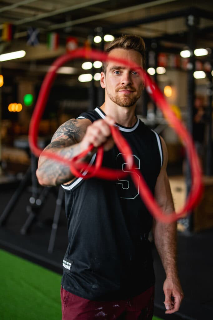 Ein bärtiger Mann mit tätowierten Armen hält ein rotes Widerstandsband in die Kamera, während er in einem Fitnessstudio steht.
