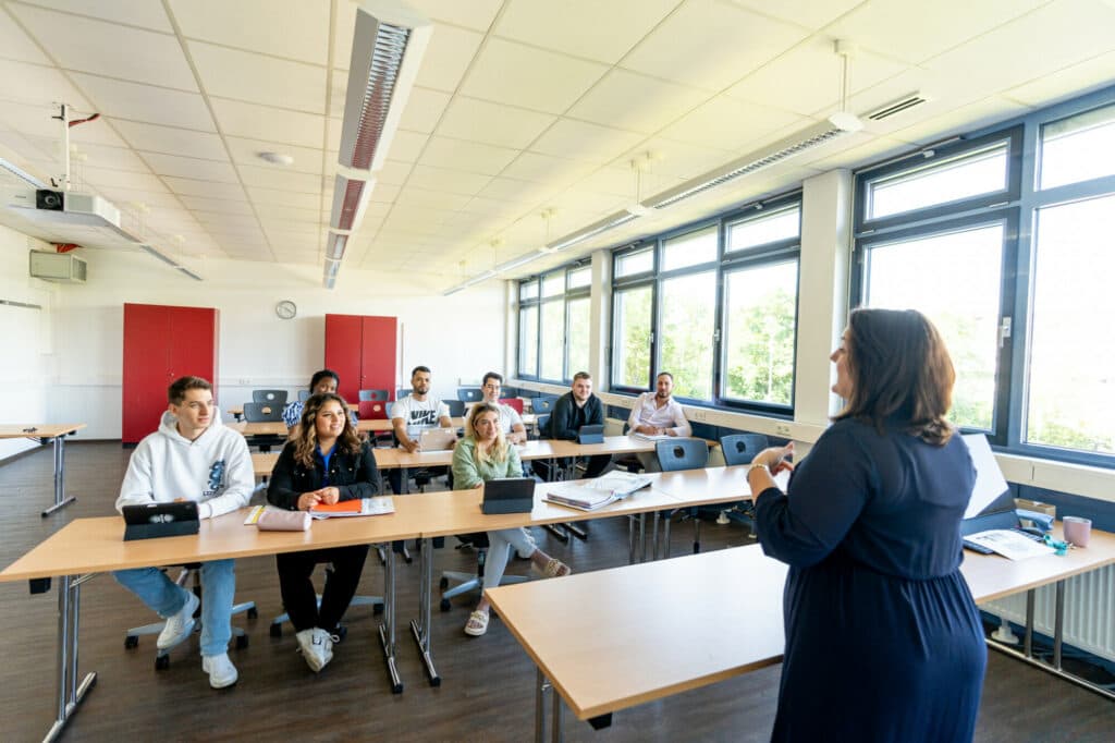 Eine Dozentin steht vor einer Gruppe von Studierenden in einem hellen Seminarraum und hält einen Vortrag.