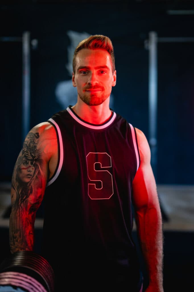 Ein muskulöser Mann mit tätowiertem Arm trägt ein dunkles Tanktop mit einem S-Logo und wird von rotem Licht angestrahlt.