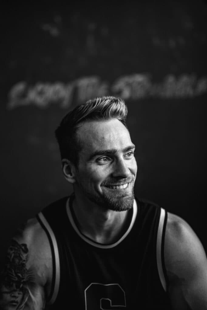 Ein schwarzweißes Porträt eines lächelnden Mannes mit Bart und tätowiertem Oberarm, der ein Sporttrikot trägt und vor einem dunklen, unscharfen Hintergrund steht.