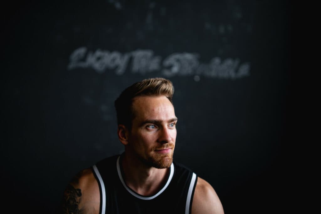 Ein Mann mit kurzem Haar und Bart trägt ein schwarzes Sportshirt und blickt nachdenklich zur Seite vor einer dunklen Wand mit weißem Schriftzug.