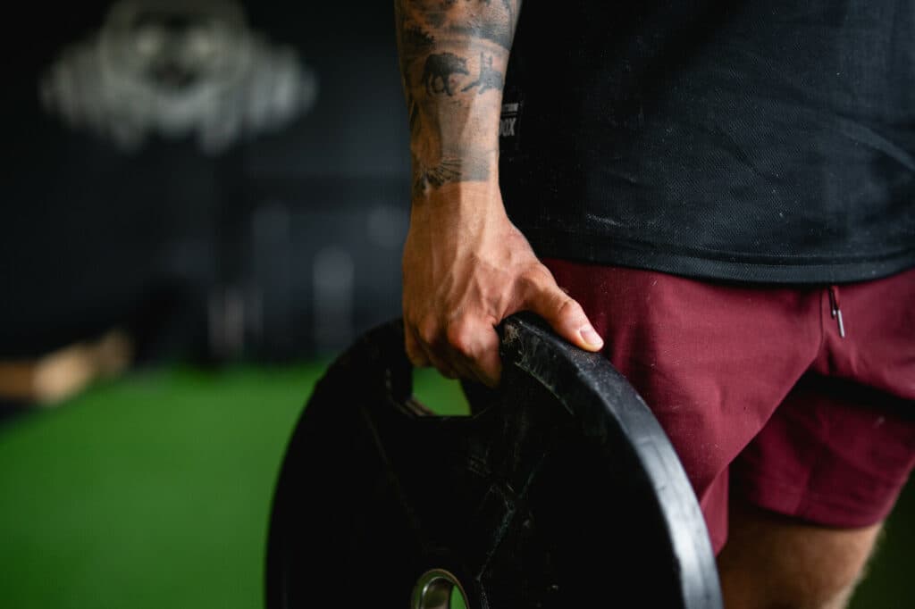 Eine tätowierte Hand hält eine schwere schwarze Hantelscheibe in einem Fitnessstudio mit grünem Boden.